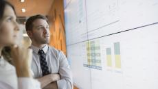 Two people standing up and looking at a large screen with graphs on it 