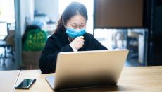 Person sitting at a desk on their laptop wearing a mask while they cough