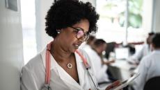 A doctor looking a tablet while a meeting occurs in the background