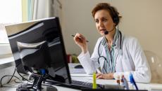 Healthcare provider sitting at a desk wearing a lab coat and headphones while talking to someone on a computer