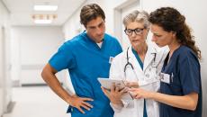 Three healthcare providers standing in the hallway of a clinical building looking at a tablet together