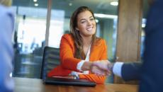 Person in an orange blazer sitting at a desk shaking hands with someone on the other side of the desk