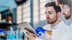 Two people in a laboratory looking at a tablet with beakers behind them