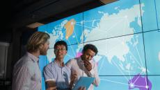 Three people standing in front of a board showing the world with lines connecting countries