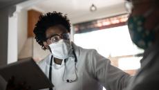 Healthcare provider wearing a mask, a lab coat and a stethoscope around their neck talking to a patient in their home