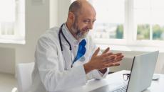 Healthcare provider with a lab coat and stethoscope sitting at a desk speaking with someone on a computer