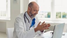 Healthcare provider wearing a lab coat and stethoscope while sitting at a desk and talking to someone on a computer