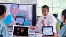 Healthcare team sitting around a table with laptops on it while talking to another healthcare provider on a TV screen